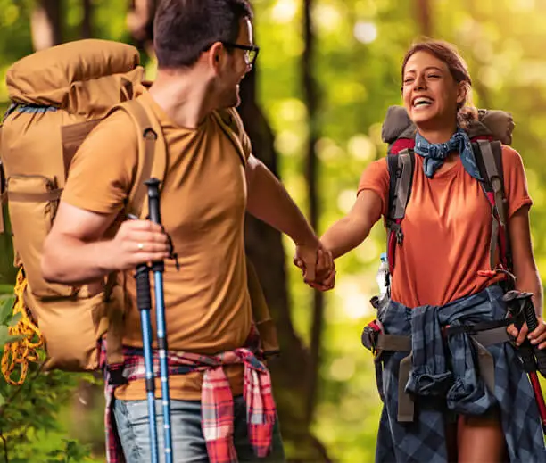 Šťastný pár na túře v lese, nesoucí batohy a držící se za ruce, užívající si přírodu a outdoorové dobrodružství.